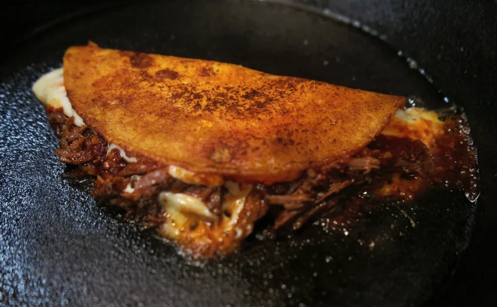 Quesabirria sizzling in a hot cast iron skillet, this helps the cheese melt and the tortilla turn out toasted. 